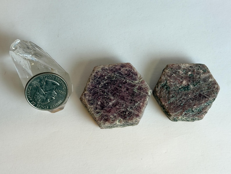 K&ouml;nnte beinhalten: Zwei hexagonale Rubin-in-Zoisit-Kristalle mit gesprenkelten dunkelroten und dunkelgr&uuml;nen Texturen, platziert neben einer klaren Quarzspitze und einer M&uuml;nze als Gr&ouml;&szlig;envergleich, die nat&uuml;rliche Mineralformationen und Kristallstrukturen zeigen.