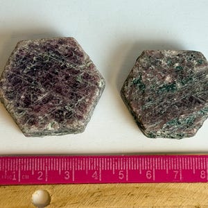 K&ouml;nnte beinhalten: Zwei hexagonale Rubin-in-Zoisit-Kristalle mit dunkelroten und dunkelgr&uuml;nen gesprenkelten Mustern. Ein rosa Lineal liegt unter den Steinen und zeigt, dass der linke Kristall etwa 3,8 cm und der rechte Kristall 3,2 cm breit ist.