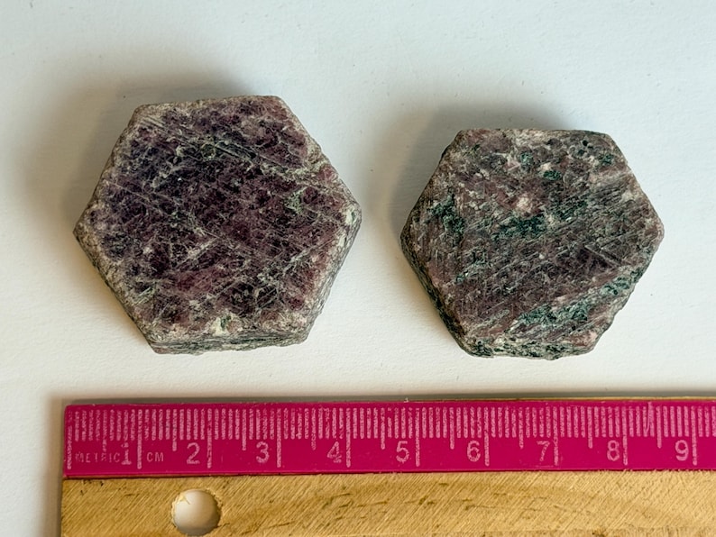 K&ouml;nnte beinhalten: Zwei hexagonale Rubin-in-Zoisit-Kristalle mit dunkelroten und dunkelgr&uuml;nen gesprenkelten Mustern. Ein rosa Lineal liegt unter den Steinen und zeigt, dass der linke Kristall etwa 3,8 cm und der rechte Kristall 3,2 cm breit ist.