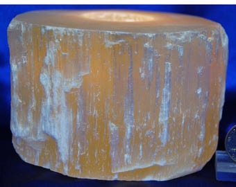 Op de afbeelding: Een cilindrische, amberkleurige selenietkristallen kandelaar. De natuursteen heeft een ruw, getextureerd oppervlak met verticale strepen. De bovenkant heeft een cirkelvormige inkeping voor een kaars. Het kristal is ongeveer 10 cm in diameter en 13 cm hoog.