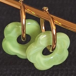 May include: A pair of gold hoop earrings with green flower-shaped charms. The charms have a swirl pattern and a glossy finish.
