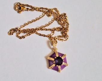 May include: A gold-colored necklace with a hexagonal purple pendant. The pendant has a dark purple center stone and is accented with gold-colored details and small clear stones. The chain is delicate and has a lobster clasp.