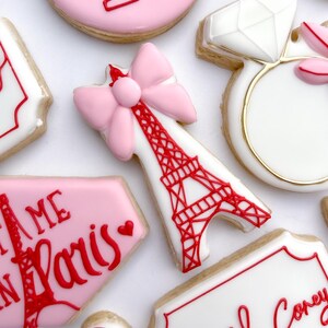 Könnte beinhalten: Ein Set dekorierter Zuckerkekse mit Pariser Thema. Die Kekse zeigen eine rosafarbene Schleife, den Eiffelturm, einen Ring mit einem Diamanten und den Text "Ich liebe Paris".