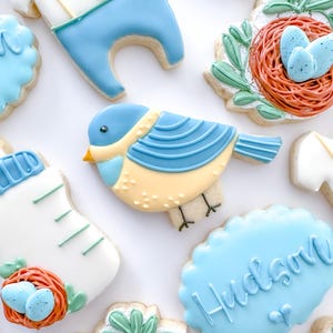 May include: A set of blue and yellow baby shower cookies decorated with a bird, a baby bottle, a nest with eggs, and a cookie with the name "Hudson" on it.