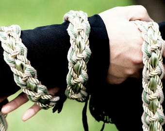 May include: A braided cord accessory featuring intertwined beige and olive green rope with a central satin ribbon. The design includes multiple loops and long, dangling ends finished with smooth olive green ribbon tassels.