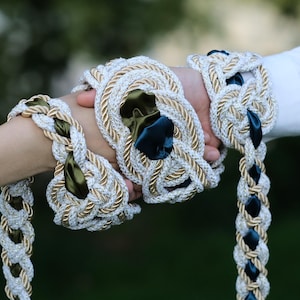 Puede incluir: Brazaletes trenzados de cuerda blanca y dorada con detalles en azul verdoso oscuro. Los brazaletes están intrincadamente tejidos y presentan detalles de cinta de raso. La imagen sugiere un conjunto de accesorios.