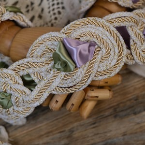 Puede incluir: Primer plano de un adorno trenzado decorativo con base color crema, detalles dorados y pequeños acentos de cinta de raso en tonos verdes y lavanda. El adorno está anudado en un patrón decorativo.