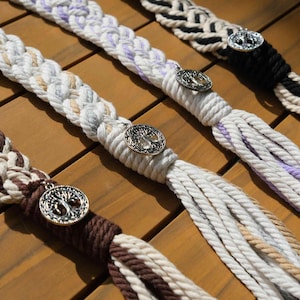 Peut inclure: Gros plan sur quatre pompons en corde tressée de différentes couleurs : marron, blanc, lavande et noir. Chaque pompon est orné d'un charm argenté décoratif avec un motif d'arbre. Les pompons sont disposés sur une surface en bois.