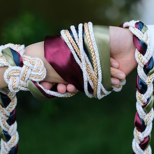 May include: A close-up of a unity braid, a wedding ceremony item. The braid is made of white, gold, burgundy, green, and navy blue ribbons. The braid is wrapped around two hands, symbolizing unity.