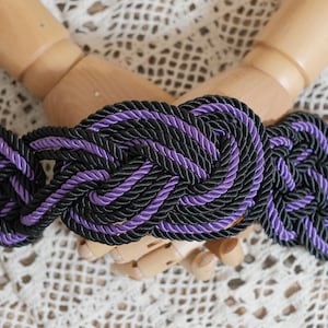 Könnte beinhalten: Ein geflochtener Gürtel aus schwarzem und violettem Seil mit einem Knotendesign.