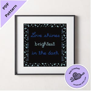 May include: A black and white cross stitch pattern with blue text that reads "Love shines brightest in the dark." The pattern is framed in a black frame and has a blue and white border.