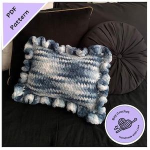 Puede incluir: Una almohada de punto de crochet a rayas azules y blancas con un borde rizado. La almohada está sobre un sofá negro con una almohada redonda negra y una almohada negra con ribete dorado. La imagen también incluye un círculo morado con el texto "Bay Crochet Hecho a mano con amor" y un corazón de crochet.
