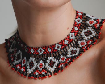 May include: A wide choker necklace crafted from small glass beads in a repeating geometric pattern. The design features a colour palette of black, white, and red, with a scalloped lower edge created by dangling bead fringe along the bottom border.