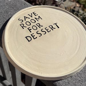 May include: A round, off-white ceramic plate with a slightly raised rim. The center of the plate features the black, stamped text "SAVE ROOM FOR DESSERT" in a simple, sans-serif font. The surface has visible circular brush strokes.