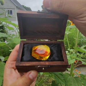 May include: A dark brown wooden box with a hinged lid, containing a faceted, translucent yellow and orange resin stone resting on a black fabric lining. The box features a metal latch on the front and is held open to display the stone inside.