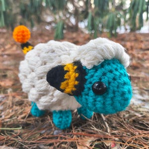 May include: A handmade crochet sheep with a white body, teal face and legs, and a black and yellow striped tail. The sheep has a fluffy white head and black button eyes. The background is blurred with brown leaves and green foliage.