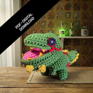 May include: A handmade green crochet dinosaur with a pink mouth, yellow teeth, and accents. The dinosaur has a large blue button eye and red ribbon details. The image includes the text "PDF • DIGITAL DOWNLOAD".