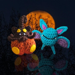 May include: Two crocheted Halloween-themed figures. One is an orange pumpkin with a brown top and yellow accents. The other is a teal bat with purple and pink accents. The background features a full orange moon and silhouettes of trees.