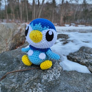 May include: A handmade crochet Piplup plushie. The blue, white, and yellow penguin-like character has black eyes and a blue scarf. It is sitting on a gray rock with a snowy background and trees in the distance.
