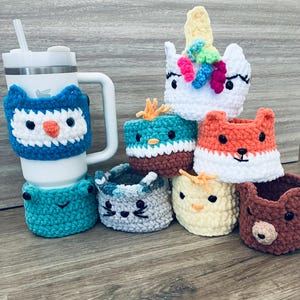May include: A collection of handmade crochet cup cozies in various animal designs, including an owl, frog, cat, fox, bear, and unicorn. The cozies are in bright colors and are designed to fit around a cup or tumbler. The owl cozy is on a white tumbler.