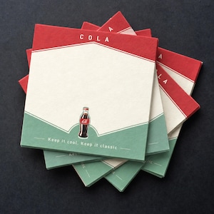 May include: A stack of square paper coasters featuring a red top section with the word COLA, a large cream-coloured centre, and a sage green bottom section. A small illustration of a glass bottle is centred, with the text Keep it cool. Keep it classic.