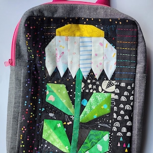 Könnte beinhalten: Eine schwarze Reißverschlusstasche mit einem rosa Griff und einem bunten Patchwork-Blumenmuster. Die Blume besteht aus verschiedenen Stoffen und hat eine gelbe Mitte, weiße Blütenblätter und grüne Blätter.