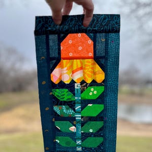 May include: A teal fabric block with a patchwork design featuring a flower with orange, yellow, and green fabric pieces. The fabric block has the text "RS & HINT 6" printed on it.
