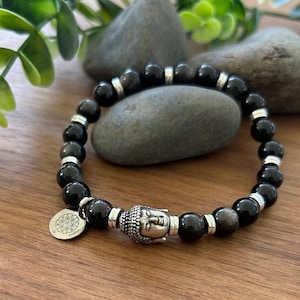 May include: Black beaded bracelet with a silver Buddha head charm and a silver charm with a flower of life design. The bracelet features black beads with silver accents and is displayed on a wooden surface.