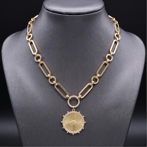 May include: A gold-toned chain necklace featuring elongated oval links connected by smaller circular rings. The necklace includes a circular pendant with a sunburst pattern, accented with small clear stones and a single central stone.