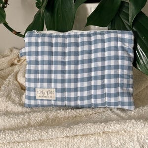 May include: A rectangular quilted pouch featuring a blue and white gingham check pattern. The pouch has a top zip closure, a small metal side loop, and a rectangular fabric label on the front that reads Pretty Petal DESIGNS CO.