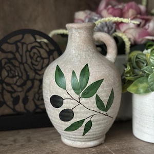 May include: A ceramic olive oil jug with a handle, painted with green olive branches and black olives. The jug has a textured, off-white glaze.