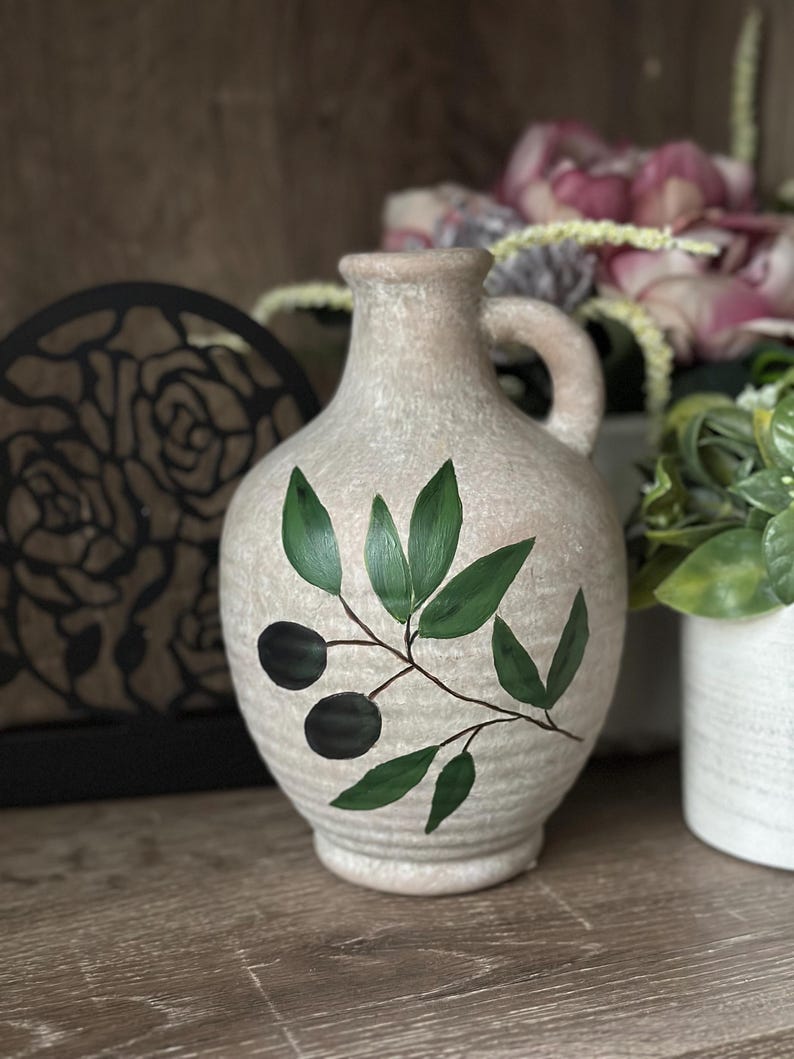 May include: A ceramic olive oil jug with a handle, painted with green olive branches and black olives. The jug has a textured, off-white glaze.