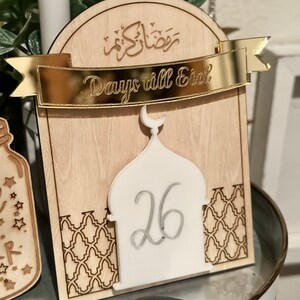 May include: A wooden countdown calendar for Ramadan with a gold banner that says "Days till Eid". The calendar features a white mosque with the number 26 inside. The background is a light wood with a decorative pattern.