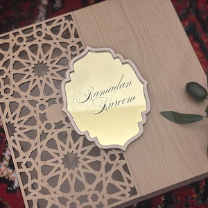 May include: A wooden box with a laser-cut geometric design on the left side and a light wood finish on the right. A gold-colored plaque with the words "Ramadan Kareem" is on top. A sprig of green leaves and an olive are on the right.