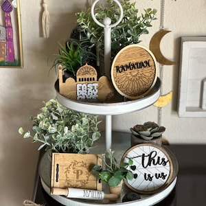 May include: A two-tiered, white metal tray with decorative items. The top tier holds greenery and a wooden piece with the word "Ramadan." The bottom tier has a wooden box, a rolling pin with text, and a round sign that says "this is us."