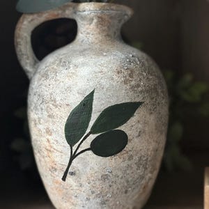 May include: A white ceramic vase with a rustic finish and a painted olive branch design. The vase has a wide mouth and a handle.