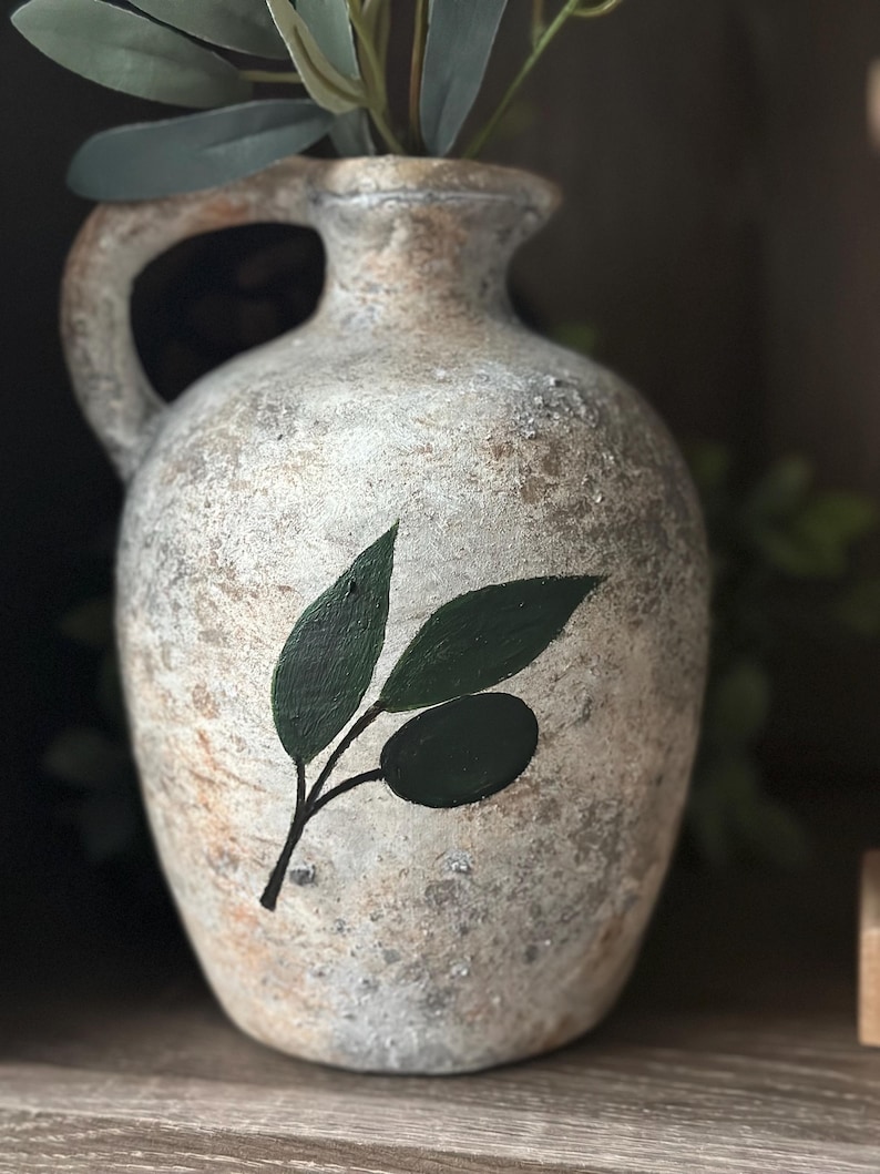 May include: A white ceramic vase with a rustic finish and a painted olive branch design. The vase has a wide mouth and a handle.