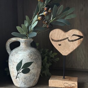 May include: A wooden heart-shaped sign with the Arabic word "حب" (meaning "love") carved into it. The sign is on a wooden stand with an infinity symbol carved into it. A vase with an olive branch design is in the background.