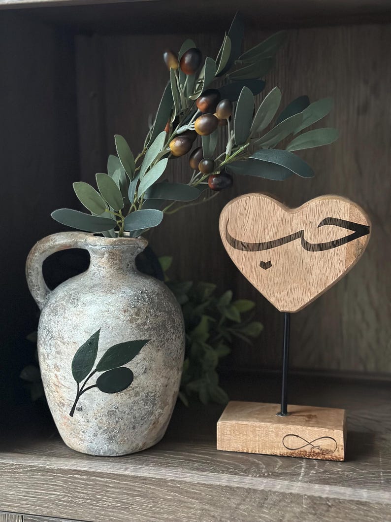 May include: A wooden heart-shaped sign with the Arabic word "حب" (meaning "love") carved into it. The sign is on a wooden stand with an infinity symbol carved into it. A vase with an olive branch design is in the background.