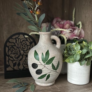 May include: A ceramic vase with a light brown glaze and a painted olive branch design. The vase has a wide mouth and a narrow neck, and it is decorated with a green olive branch with black olives. The vase is sitting on a wooden surface.