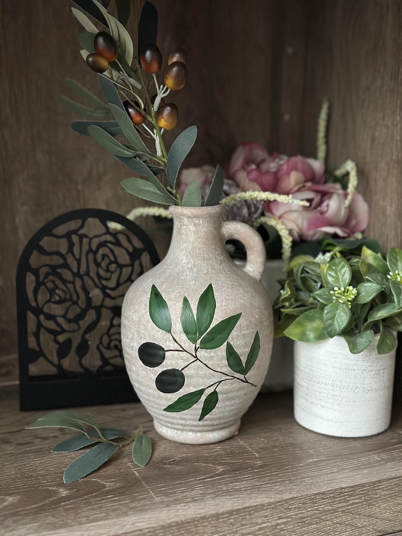 May include: A ceramic vase with a light brown glaze and a painted olive branch design. The vase has a wide mouth and a narrow neck, and it is decorated with a green olive branch with black olives. The vase is sitting on a wooden surface.