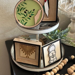 May include: A tiered tray with four wooden framed wall decor pieces. The top piece is a round olive branch design with a green background and a wood cutout of the Palestinian territory. The second piece is a rectangular wood cutout of the Palestinian territory with the Palestinian flag colors. The third piece is a square with a traditional Palestinian embroidery design. The fourth piece is a square with a cartoon illustration of a person praying. The tray is decorated with wooden beads and a black tassel.