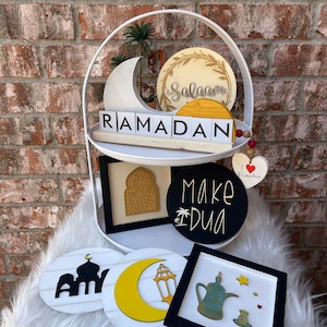 May include: A white two-tiered serving tray with Ramadan-themed decorations. The tray holds wooden cutouts of a crescent moon, a mosque, and the word "RAMADAN". Other decorations include a circle with the word "Salaam" and a black circle with the words "Make Dua".