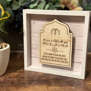 May include: A wooden plaque with an Islamic prayer in Arabic and English. The plaque is framed in a white frame and has a light wood background. The prayer reads "Bismillah walajna, wa bismillah kharajna, wa ala rabbana tawakkalna" in Arabic and "In the name of Allah we enter, and in the name of Allah we leave, and upon our Lord, we place our trust" in English.
