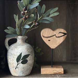 May include: A wooden heart-shaped sign with the Arabic word for love carved into it. The sign is on a wooden base with an infinity symbol carved into it. A vase with a green olive branch design sits on a shelf behind the sign.