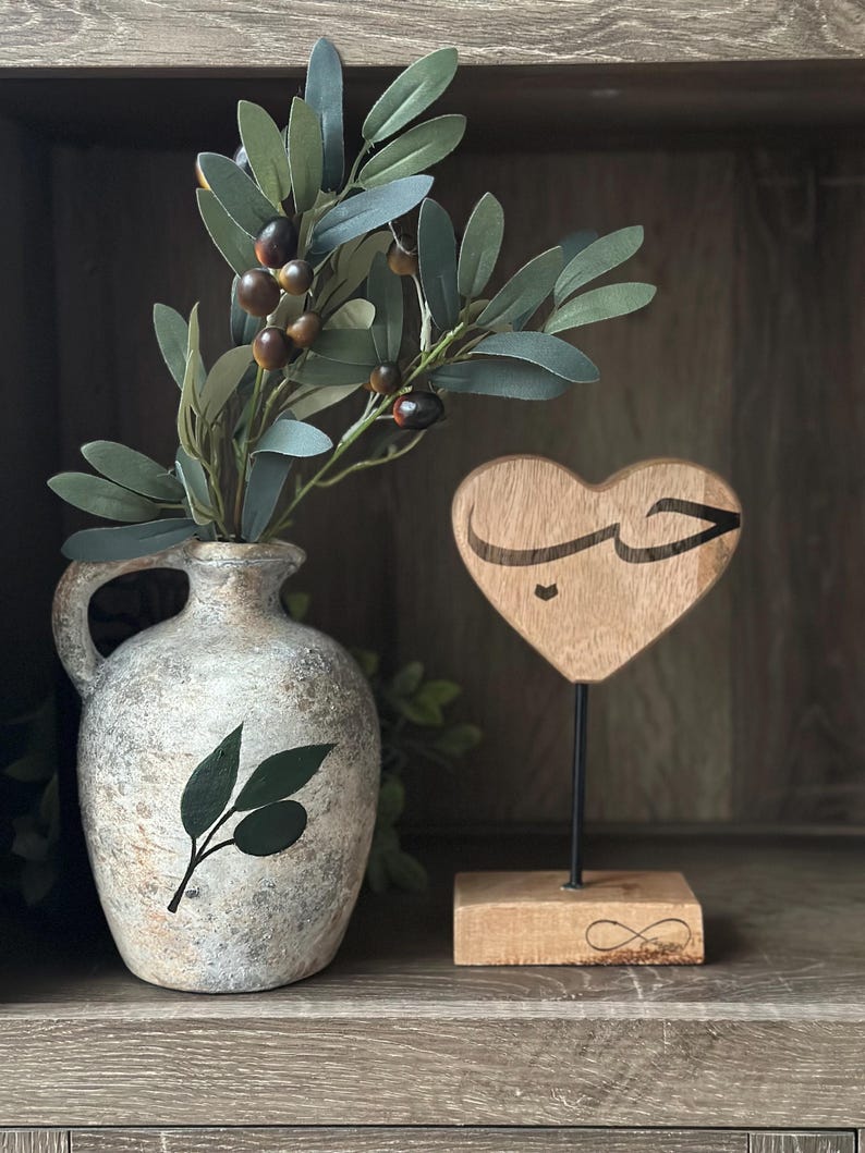 May include: A wooden heart-shaped sign with the Arabic word for love carved into it. The sign is on a wooden base with an infinity symbol carved into it. A vase with a green olive branch design sits on a shelf behind the sign.