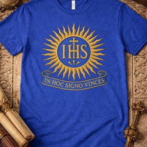 May include: Royal blue t-shirt featuring a gold emblem with the letters IHS, a cross, and the Latin phrase "IN HOC SIGNO VINCIS" within a sunburst design. The shirt is made of a soft material.
