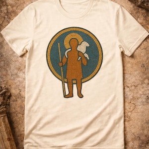 May include: Cream-colored t-shirt featuring a mosaic-style graphic of a figure holding a staff and a lamb. The figure is brown, set against a teal and gold circular background. The shirt is made of a soft material.