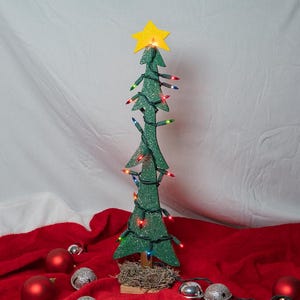May include: A flat, green, glittery wooden Christmas tree cutout featuring a yellow star topper and wrapped in a string of multicolored LED lights. The tree stands on a small, textured base, surrounded by red and silver spherical ornaments.