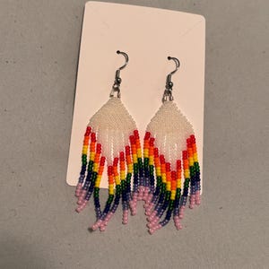 Può includere: Un paio di orecchini di perline con un design arcobaleno. Gli orecchini presentano una parte superiore di perline bianche con una frangia di perline rosse, arancioni, gialle, verdi, blu e viola. Sono appesi a ganci color argento.